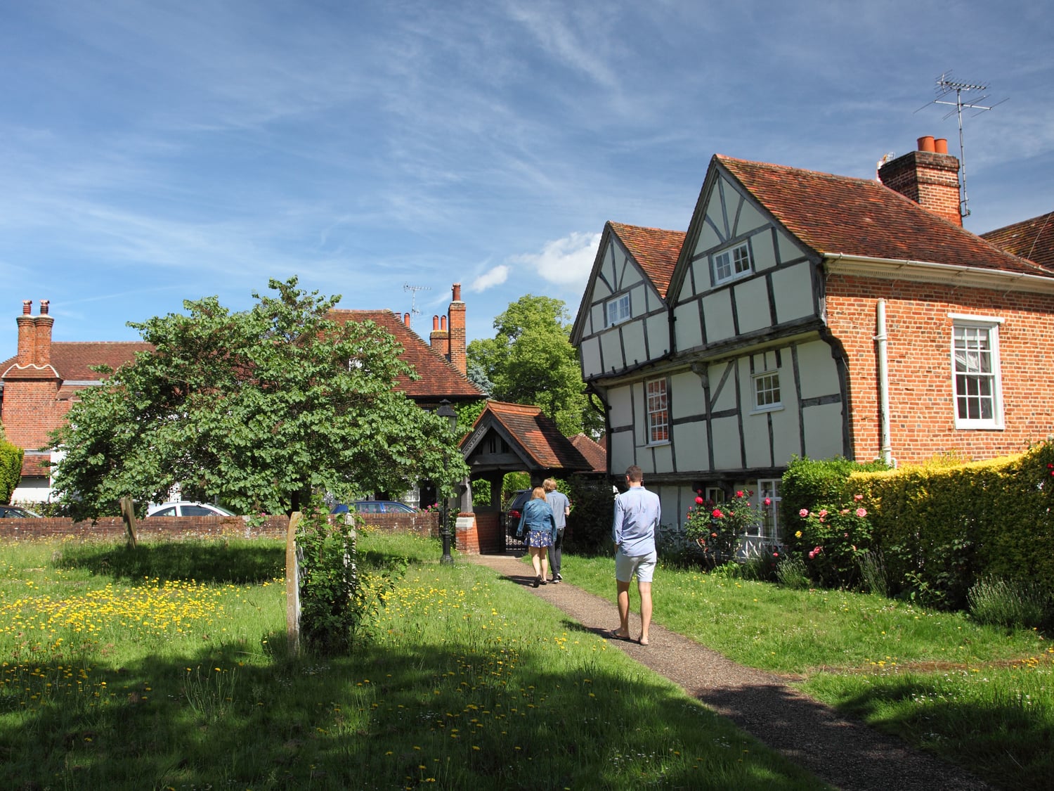 A few people walking through the grounds of St. Andrew's Church, Cobham, next to a Tudor style building.