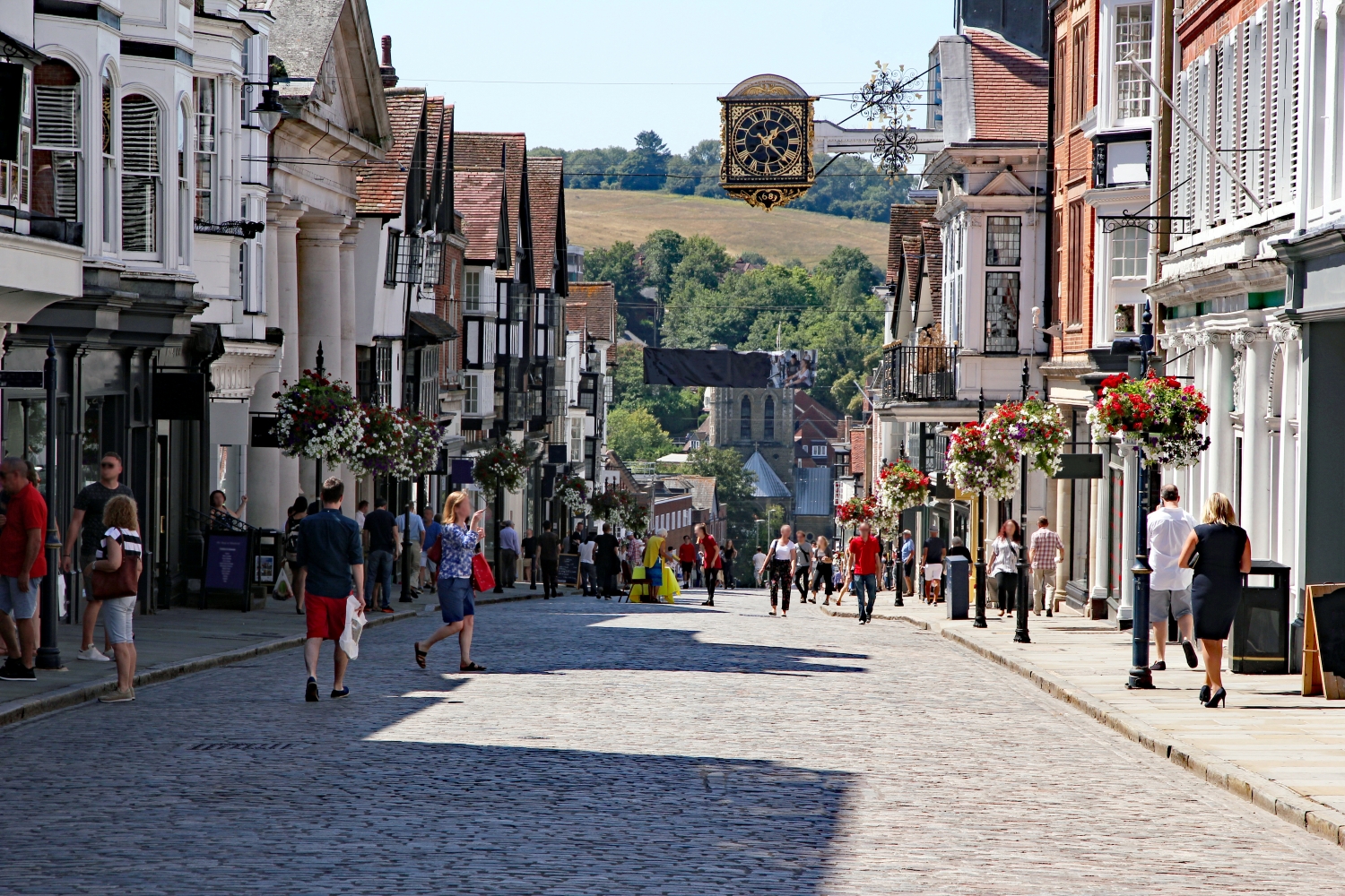 Guildford High Street with its historic cobbled road, traditional buildings, hanging flower baskets and the iconic Guildhall clock in Surrey town centre.