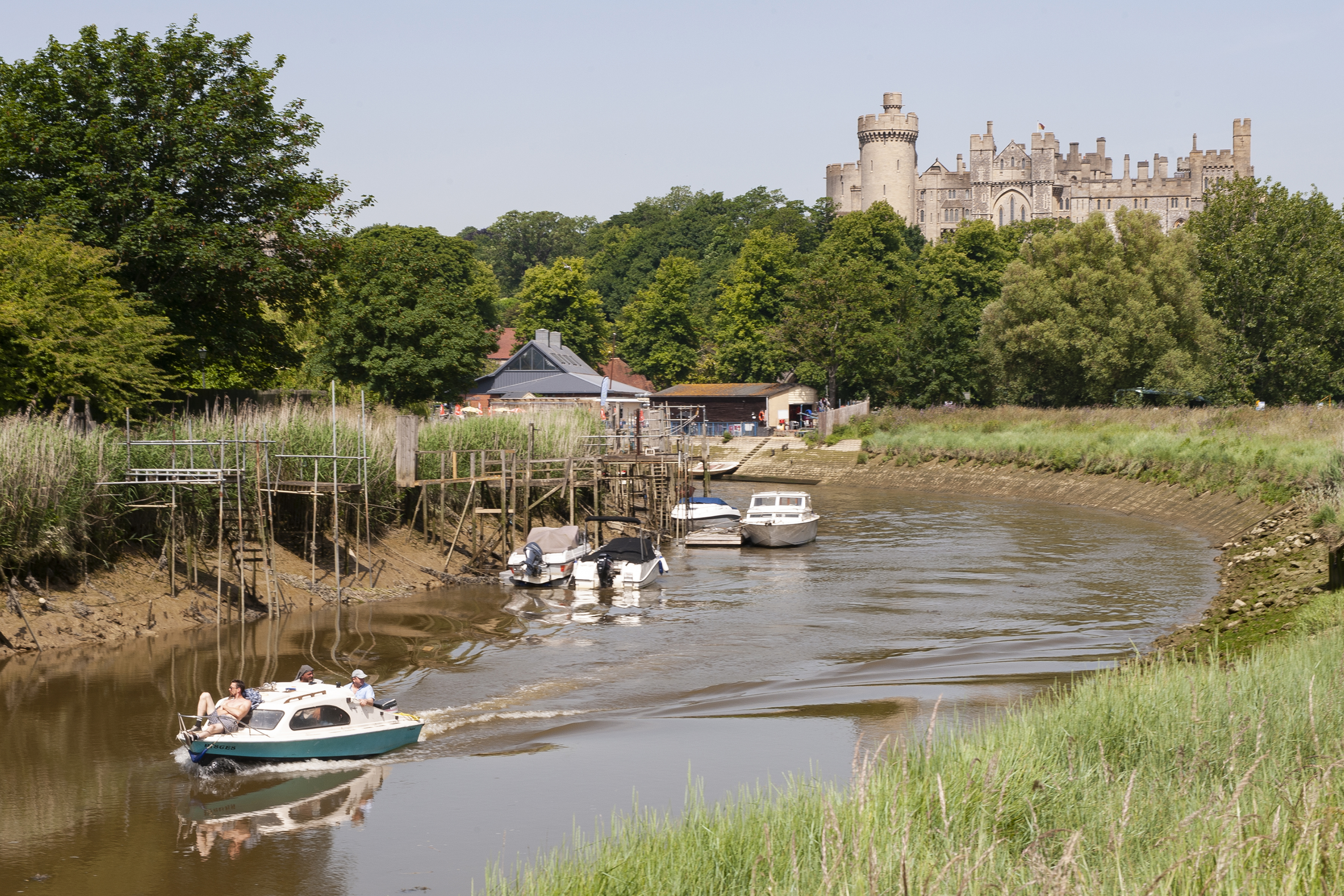Arundel Castle set back behind a tree line with the river Arun flowing in front of shot. Boats are travelling down the river.