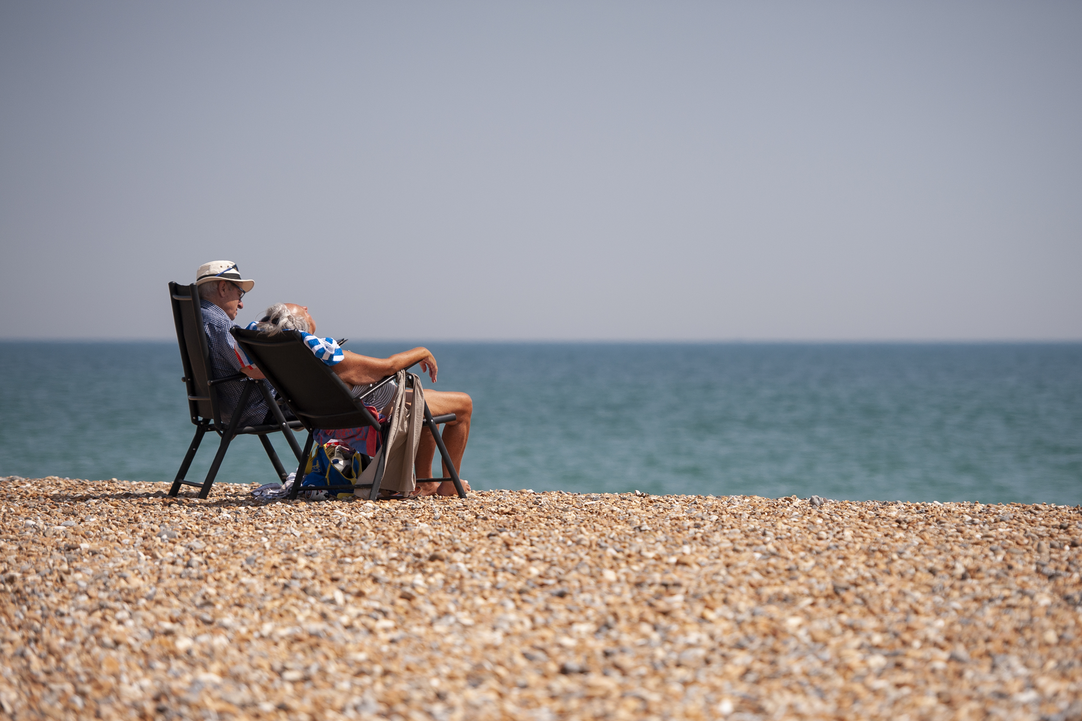 Two people sit on camping chairs tanning in the sun over a pebble beach.