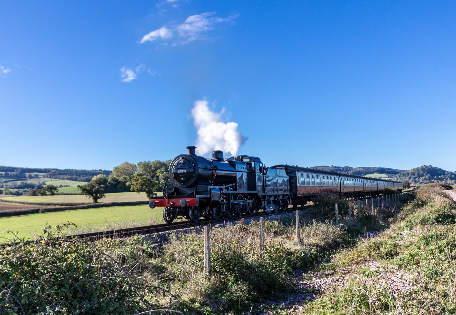 A vintage steam train and carriages on the West Somerset Railway with green fields and blue sky in the background.
