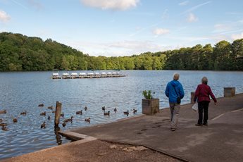 Two retirees walk alongside water populated with ducks in Boldmere Gate.