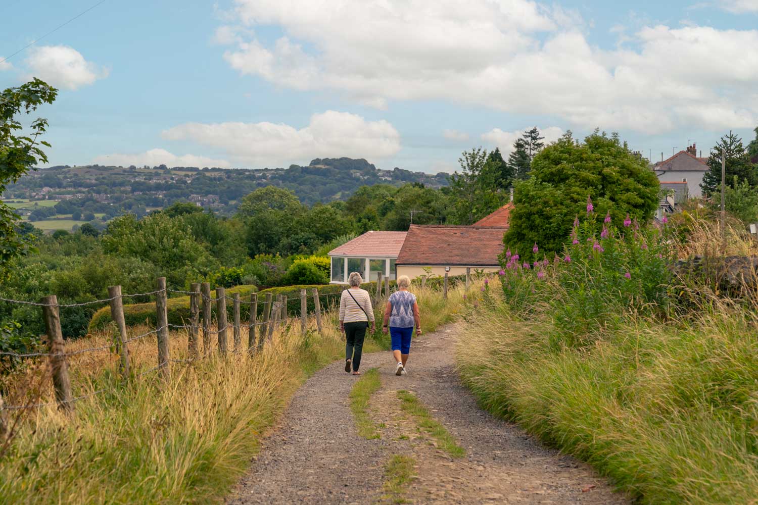 a couple people walking on a dirt path in a grassy area with a house and trees in the