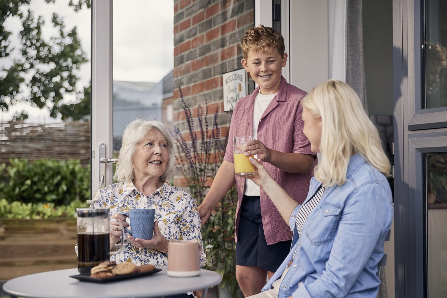 A child, their parent and a senior on the patio of a McCarthy Stone retirement property, talking whilst enjoying refreshments.