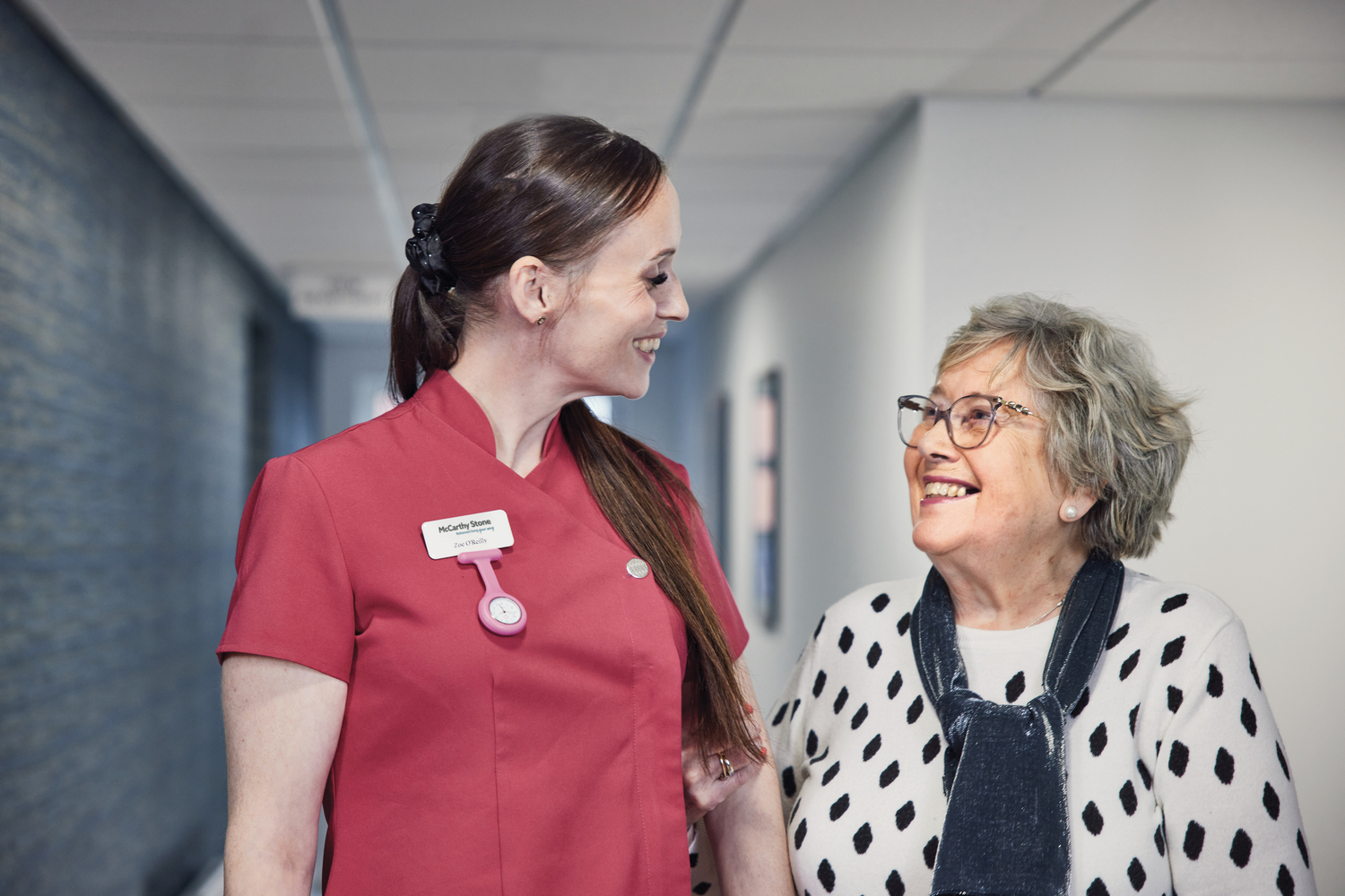 Elderly woman walking and smiling with carer in supported living corridor.