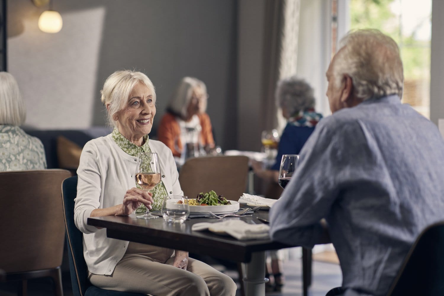 Seniors enjoying eating at the bistro at a Retirement Living PLUS development by McCarthy Stone. Comfy chairs and booths complement the contemporary interior design